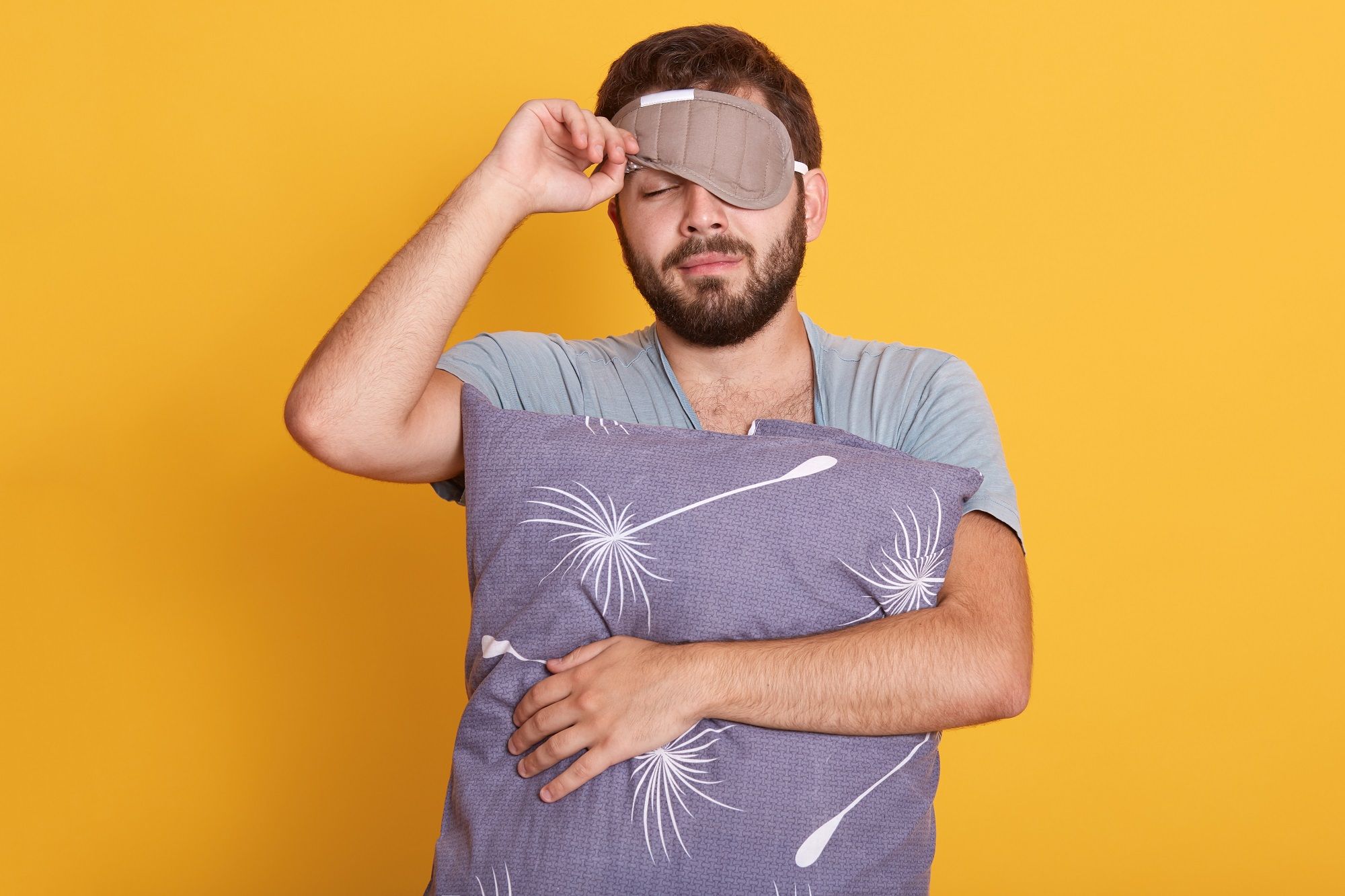 closeup portrait sleepy man with blindfold eyes holding pillow hands opens sleeping mask keeping eyes closed