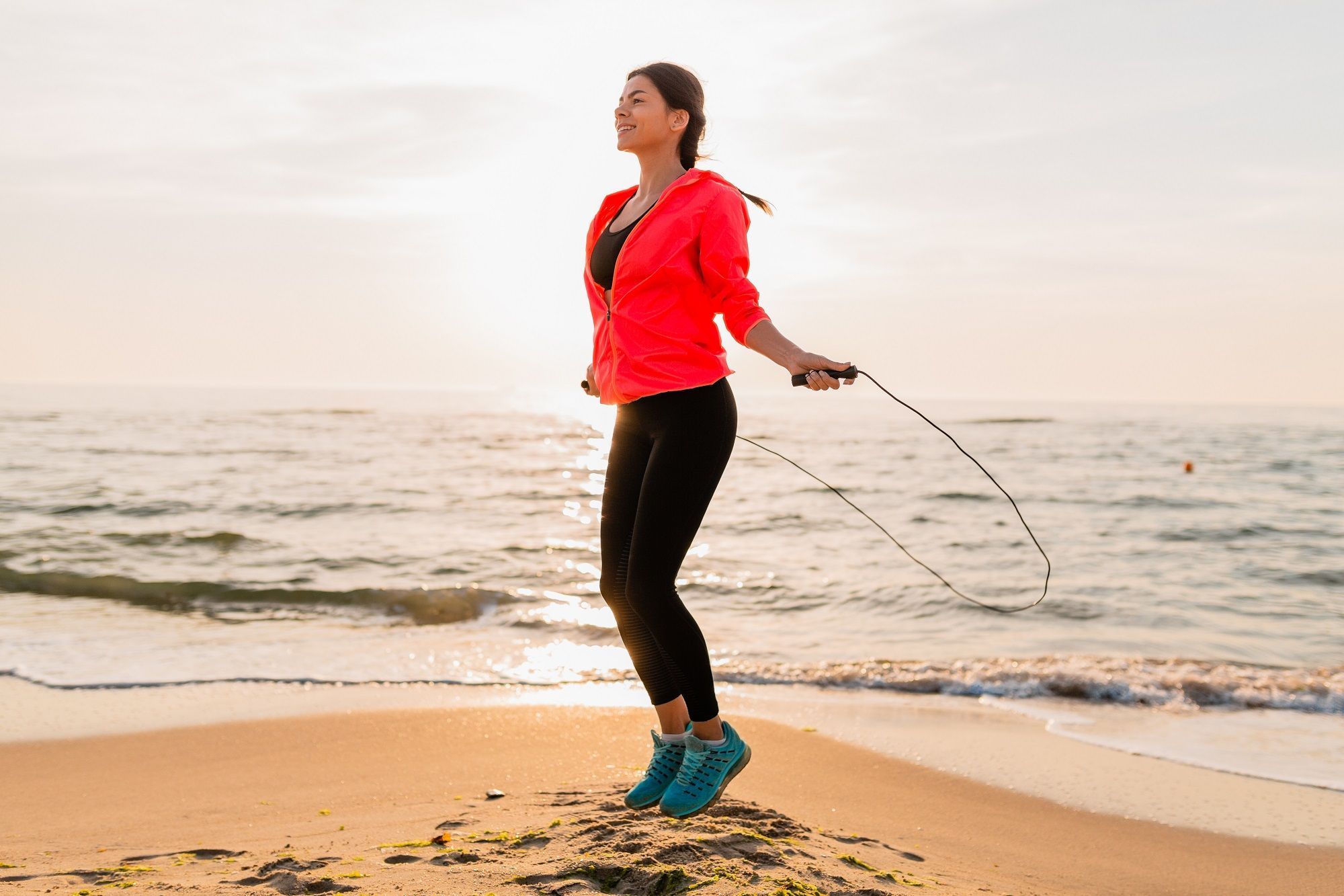 young attractive woman doing sport exercises morning sunrise sea beach sports wear healthy lifestyle listening music earphones wearing pink windbreaker jacket jumping jump rope