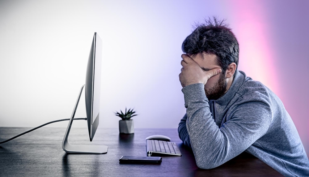 A tired man sits in front of a computer covering his face with his hands
