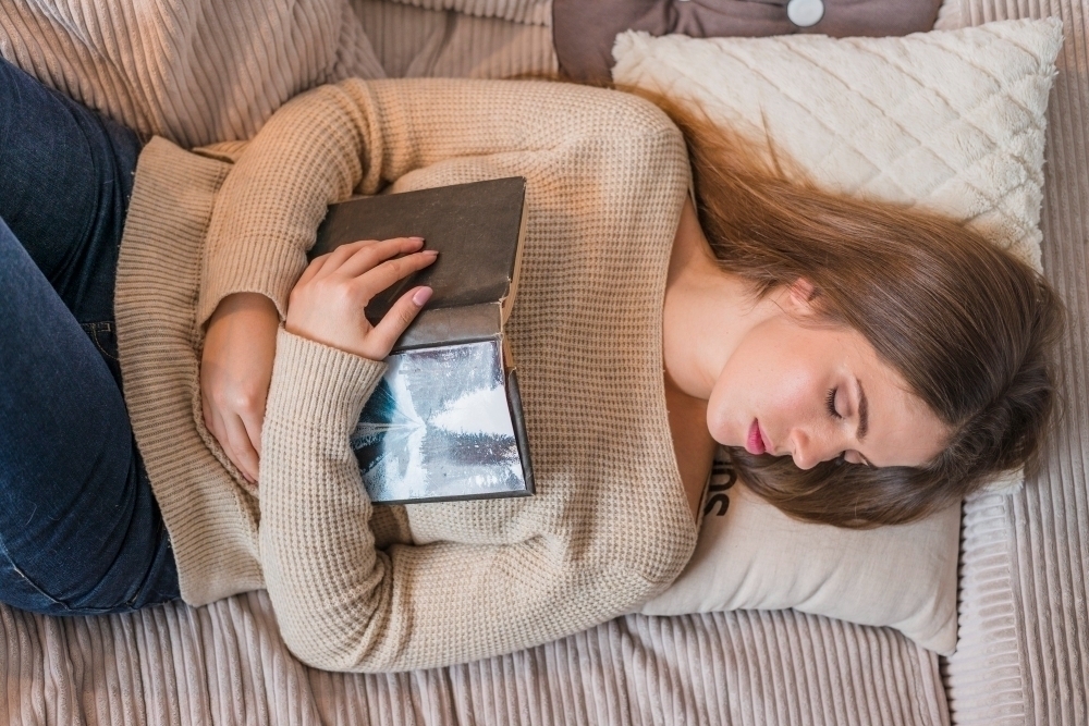 An overhead view of young woman sleeping on bed with book in hands