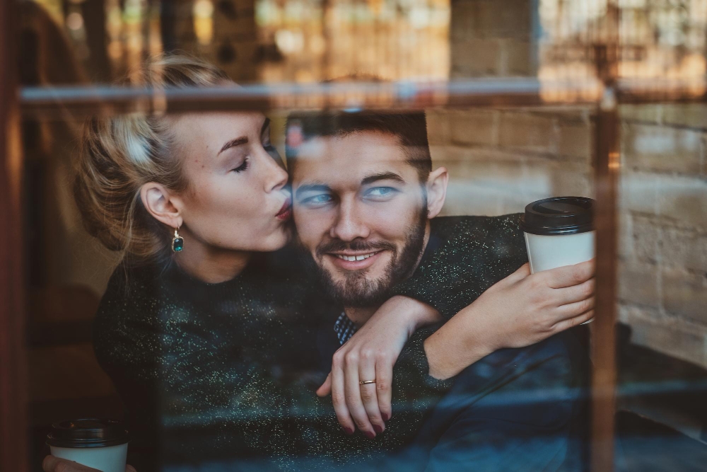 Attractive woman is kissing her man while sitting at cafe with cup of coffee.