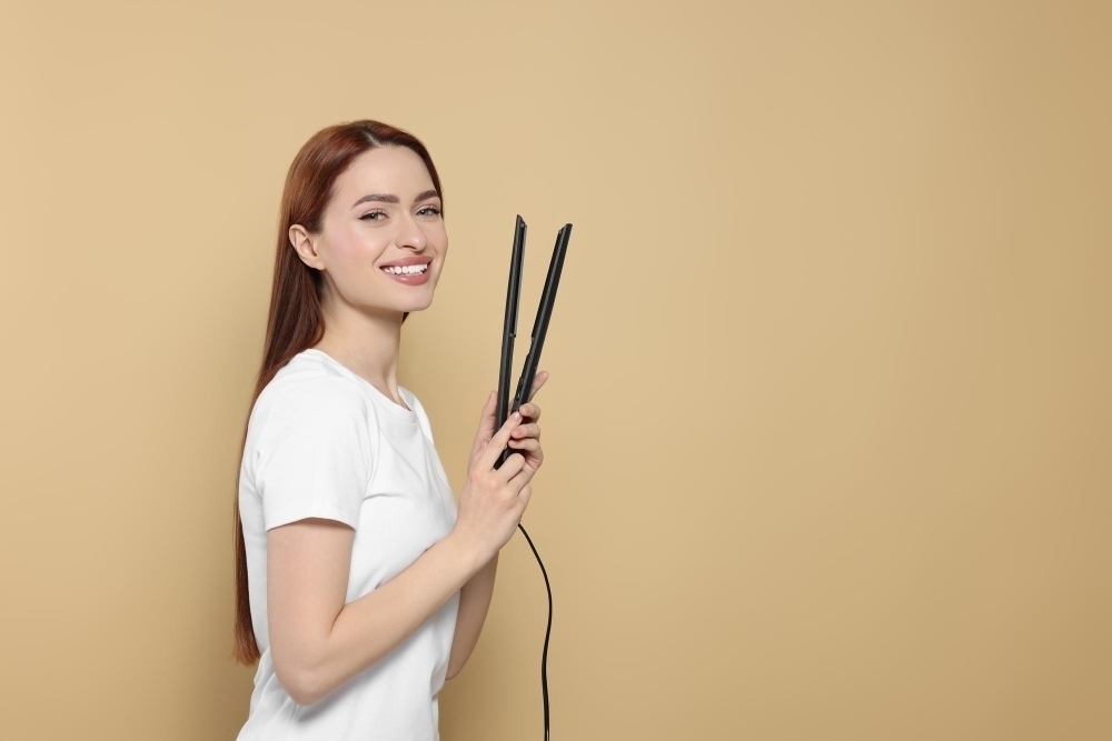 Beautiful woman with hair iron on beige background space for text