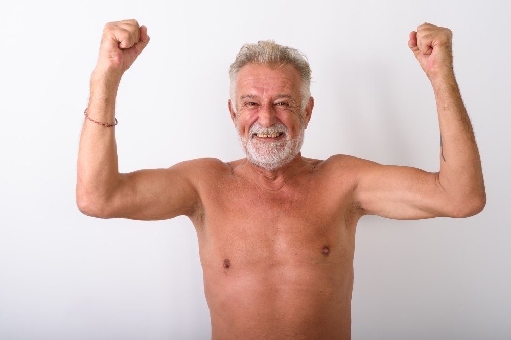 Happy senior bearded man smiling while looking excited shirtless with both arms raised on white