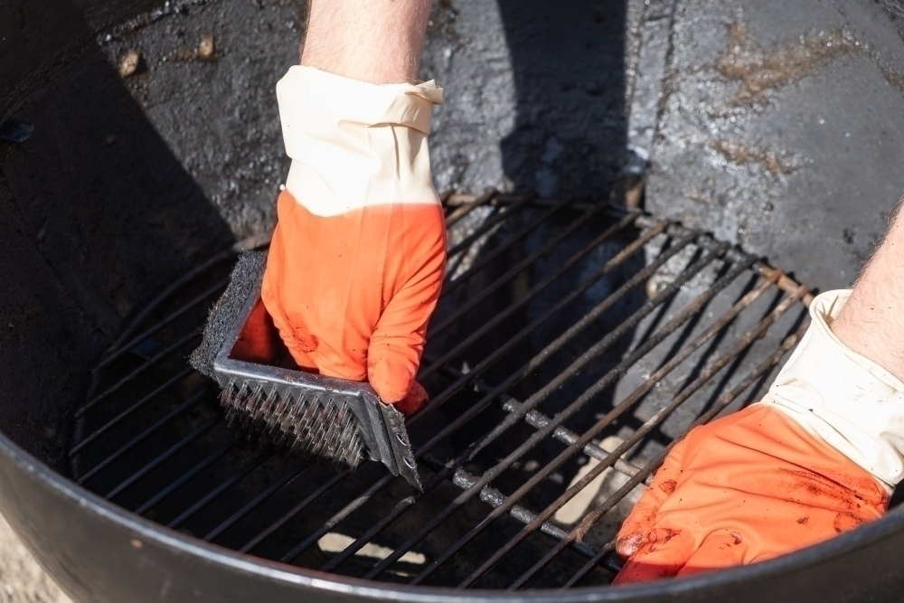 Male hand with red gloves cleans round grill with stiff brush.