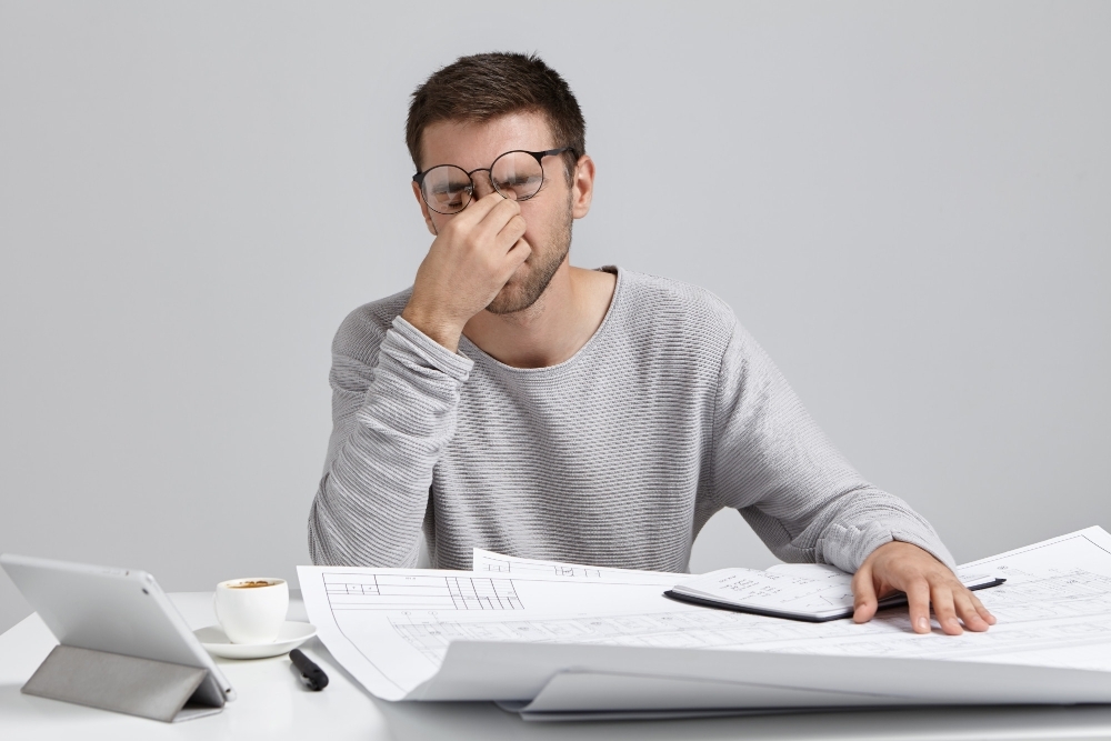 Stess overwork and deadline. Tired unshaven young architect freelancer massaging nose bridge