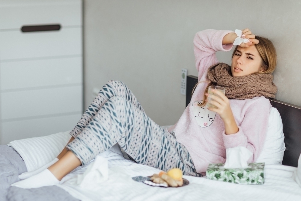 Young woman sick in bed with temperature drinks hot tea and touching her head
